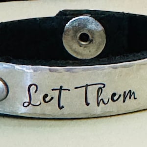 May include: Black leather bracelet with a silver metal plate that says "Let Them". The plate has a hammered texture and is secured with two silver rivets.