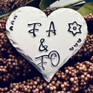 Puede incluir: Un colgante plateado en forma de corazón con las iniciales "FA & FO" estampadas. El colgante tiene una superficie texturizada y está decorado con pequeños diseños florales y curvos. El fondo es una mezcla de marrón y verde.