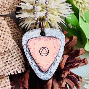 以下が含まれることがあります： レイヤードペンダントネックレス。銅色のギターピック型のペンダントには、黒い三角形が円で囲まれており、テクスチャ加工された銀色の金属にセットされています。ペンダントはシルバーのボールチェーンから吊り下げられています。