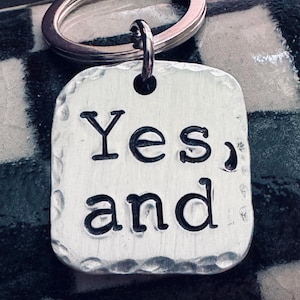 May include: A silver key chain with a rectangular tag that says "Yes, and" in a vintage font.