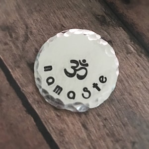 May include: A silver charm with a hammered texture featuring the Om symbol and the text "namaste" around the edge.