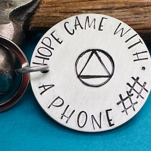 May include: A silver-colored round keychain tag with the words "HOPE CAME WITH A PHONE #" stamped around the edge. The center features a black triangle within a circle. A silver key ring is attached.