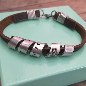 May include: A brown leather bracelet with silver metal charms. The charms are engraved with the words "Forever" and a tree design.