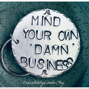 May include: Silver keychain with a round charm that says "Mind your own *damn* business".