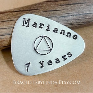 May include: Silver guitar pick with the text "Marianne 7 years" and a black triangle symbol inside a circle.