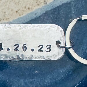 May include: Silver keychain with a hammered rectangular tag engraved with the date "11.26.23".