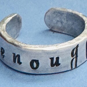 May include: A silver ring with the word "enough" engraved on it.