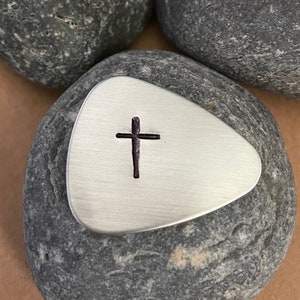 May include: A silver-toned guitar pick with a brushed metal finish featuring a small, dark, stamped cross symbol centered on the front surface. The pick has a triangular shape with rounded corners and a smooth, flat appearance.