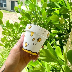 May include: White ceramic planter with a yellow and black bee design. The planter has a yellow rim and a black outline around the bees. The bees are flying and have a dotted line behind them.