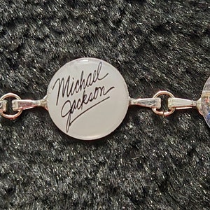May include: A silver bracelet with round medallions featuring images of Michael Jackson. The medallions have a silver border and are connected by silver chains. One medallion has the text "Michael Jackson" written in black.
