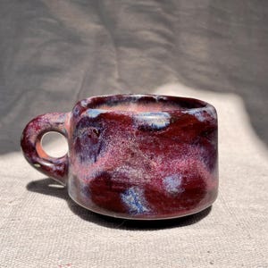 May include: A ceramic mug with a rounded shape and a small handle, featuring a glossy glaze in deep shades of burgundy and maroon, accented with swirling patches of light blue and white throughout the exterior surface of the vessel.