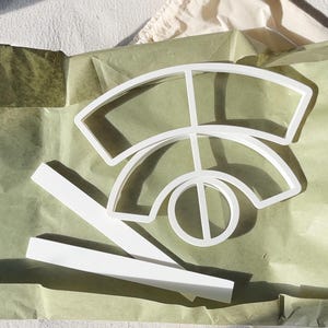 May include: White plastic cookie cutters in various geometric shapes, including a semi-circle, a circle with a line through it, and two rectangular pieces. The cutters are arranged on green tissue paper.