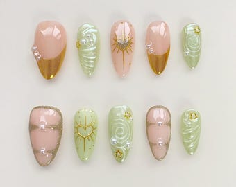 May include: Set of ten false nails with a pastel green and pink color scheme. The nails feature various gold accents, including glitter, stripes, and designs. Some nails have small pearl embellishments.