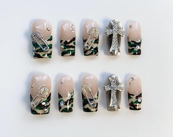 May include: Ten fake nails with a camouflage pattern. Five nails have a gold safety pin design and five nails have a silver cross design.