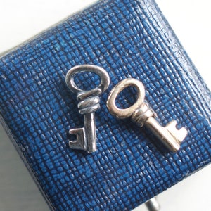May include: Two silver-colored key charms rest on a textured, dark blue box. The keys have detailed designs, and the box has a square shape. The background is a bright white surface.