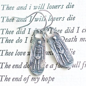 May include: Silver skeleton earrings with a textured finish. The earrings are shaped like small rectangular pendants and feature a simple skeleton design. The text "Thee did I chuse; Death must loo" is visible in the background.