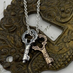 May include: A silver and gold key necklace with two antique-style keys hanging from a silver chain. The keys are detailed with intricate designs and have a worn, aged look.
