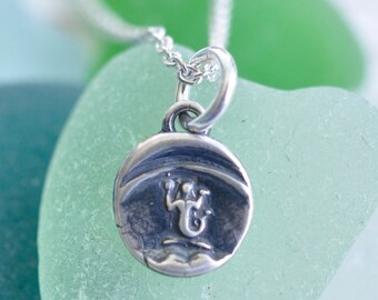 May include: A small, circular sterling silver pendant on a delicate silver chain. The pendant features an oxidized finish and a central embossed design of a stylized figure with raised arms, set against a smooth, rounded metal surface.