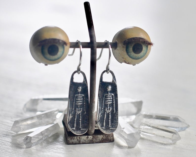 May include: Silver skeleton earrings with a silver hook closure. The earrings are hanging from a metal stand with two fake eyes on either side.