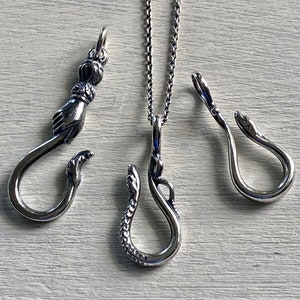 May include: Three silver pendant necklaces with different designs. The first pendant is a hand holding a hook. The second pendant is a snake with a hook. The third pendant is a snake with a hook.