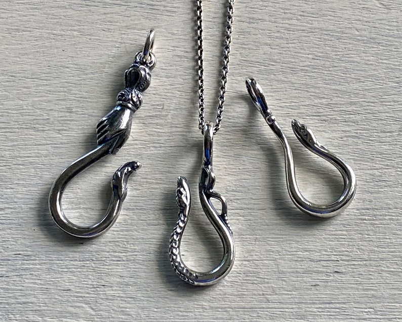 May include: Three silver pendant necklaces with different designs. The first pendant is a hand holding a hook. The second pendant is a snake with a hook. The third pendant is a snake with a hook.