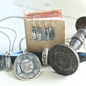 May include: A set of silver skeleton earrings, a silver skeleton ring, and two silver skeleton pendants on a chain. The jewelry is displayed on a small wooden box and a black and silver stand.