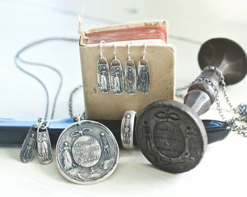 May include: A set of silver skeleton earrings, a silver skeleton ring, and two silver skeleton pendants on a chain. The jewelry is displayed on a small wooden box and a black and silver stand.