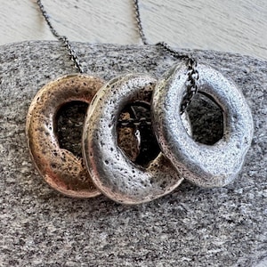 Könnte beinhalten: Drei strukturierte Metall-Anhänger-Halsketten mit kreisförmigen Designs. Ein Anhänger ist aus Bronze, einer aus Silber und einer aus einer Mischung aus Silber und Bronze. Die Halsketten werden auf einer grauen Steinoberfläche präsentiert.