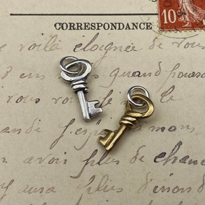 May include: Two small decorative keys, one silver-toned and the other gold-toned, rest on a vintage postcard with faded handwriting. The keys have intricate details and are likely charms or pendants.