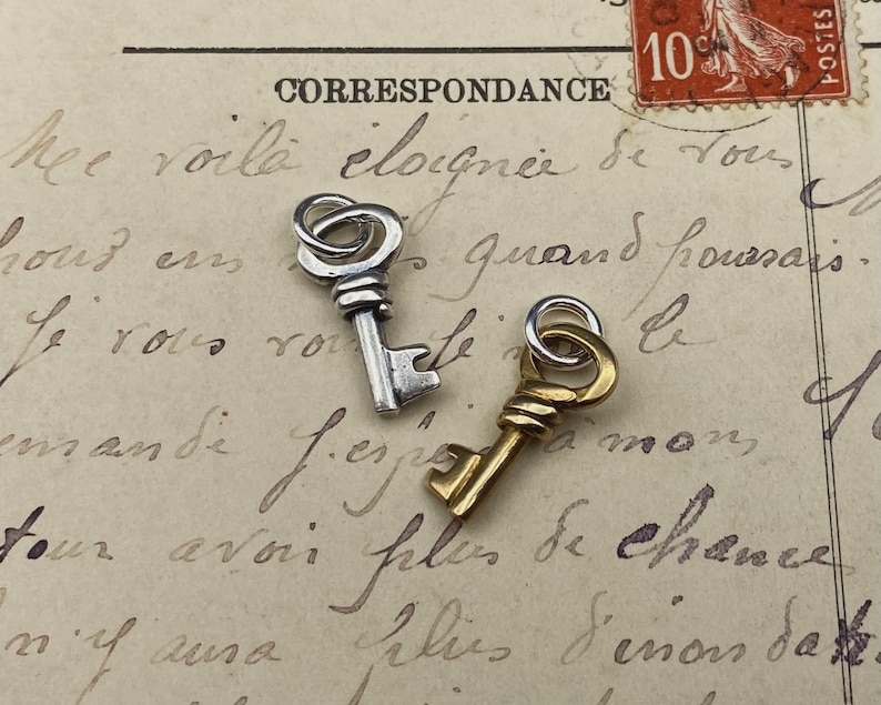May include: Two small decorative keys, one silver-toned and the other gold-toned, rest on a vintage postcard with faded handwriting. The keys have intricate details and are likely charms or pendants.