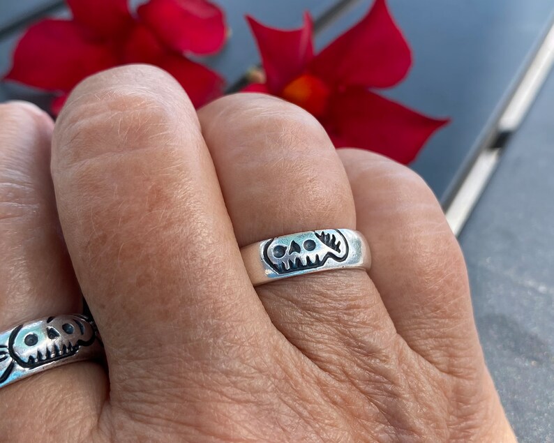 Pu&ograve; includere: Un anello d'argento con un designo a forma di teschio. L'anello &egrave; indossato su un dito.