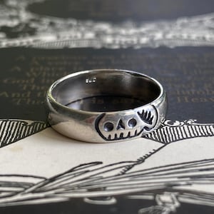 Pu&ograve; includere: Un anello d'argento con un designo a forma di teschio. L'anello &egrave; spesso e ha una fascia arrotondata.