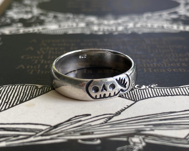 Pu&ograve; includere: Un anello d'argento con un designo a forma di teschio. L'anello &egrave; spesso e ha una fascia arrotondata.