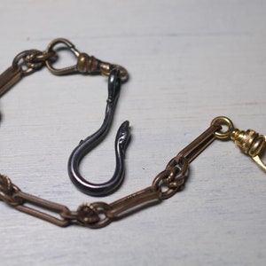 May include: A gold pocket watch chain with a black metal hook. The chain has a decorative design and a swivel clasp.