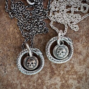 May include: Two silver necklaces with skull pendants. Each pendant is a circle with a snake wrapped around it. The snake's head is at the top of the circle and the tail is at the bottom. The skull is in the centre of the circle.