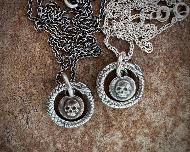 May include: Two silver necklaces with skull pendants. Each pendant is a circle with a snake wrapped around it. The snake's head is at the top of the circle and the tail is at the bottom. The skull is in the centre of the circle.
