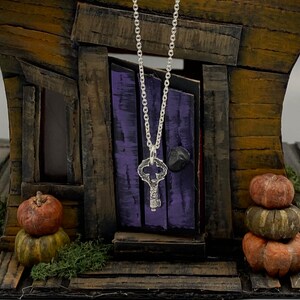 May include: A silver key pendant necklace hanging in front of a miniature wooden house with a purple door. The house is decorated with small pumpkins.