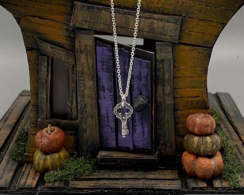 May include: A silver key pendant necklace hanging in front of a miniature wooden house with a purple door. The house is decorated with small pumpkins.