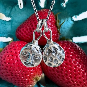 May include: A silver pendant necklace featuring two round charms. One charm has a face with closed eyes, and the other has a textured surface. The necklace hangs in front of bright red strawberries.