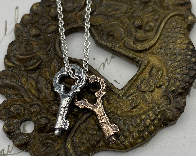 May include: A silver and gold key necklace with two antique-style keys hanging from a silver chain. The keys are detailed with intricate designs and have a worn, aged look.