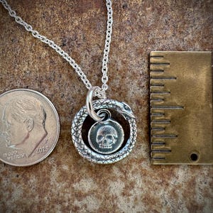 May include: A silver necklace featuring a snake charm encircling a skull pendant. The necklace is approximately 18 inches in length and is displayed alongside a dime for scale.