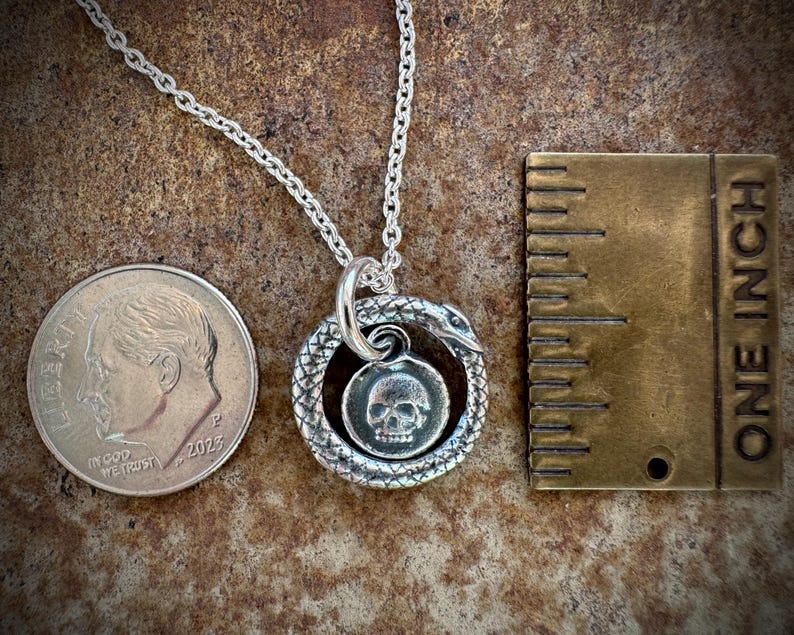 May include: A silver necklace featuring a snake charm encircling a skull pendant. The necklace is approximately 18 inches in length and is displayed alongside a dime for scale.