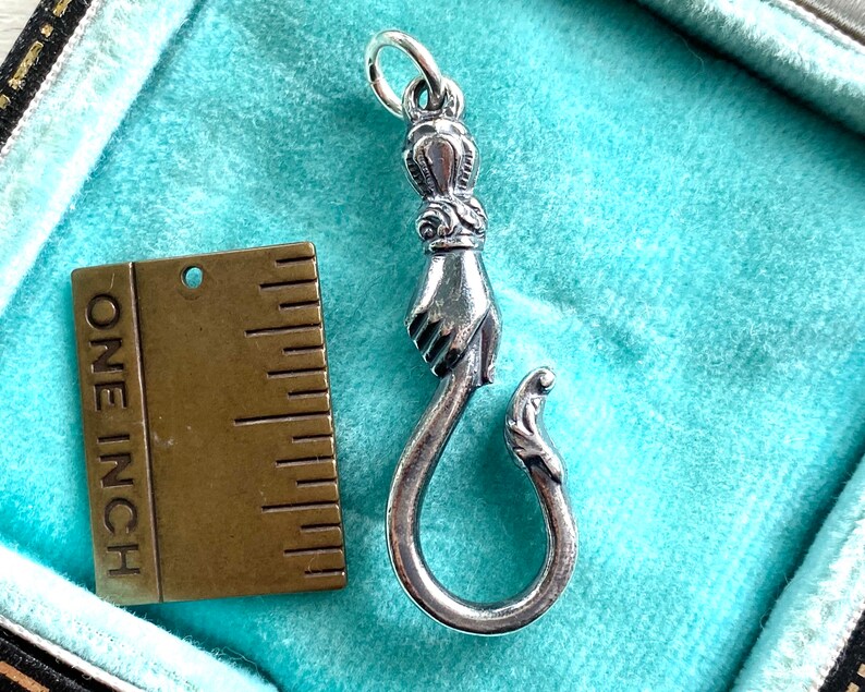May include: A silver charm in the shape of a hand holding a hook. The charm is about one inch long and is displayed on a blue background with a ruler.