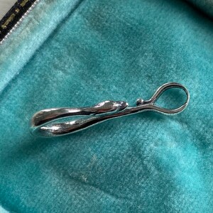 May include: A silver-toned metal clasp with a smooth, curved design. The clasp is open and appears to be a decorative element for a piece of jewelry.