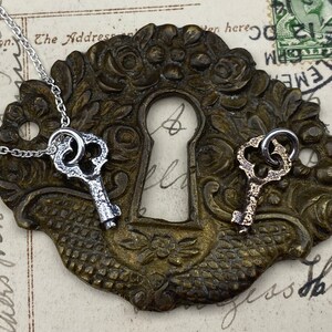May include: Two silver key charms on a chain, one with a silver finish and one with a gold finish, are displayed on a vintage brass keyhole cover.