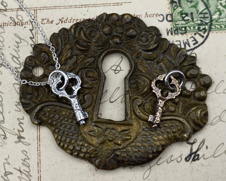 May include: Two silver key charms on a chain, one with a silver finish and one with a gold finish, are displayed on a vintage brass keyhole cover.