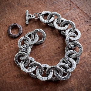 May include: A silver-toned chain link bracelet with a toggle clasp and a matching ring. The bracelet and ring have a textured, rustic appearance. The jewelry is displayed on a dark brown wooden surface.