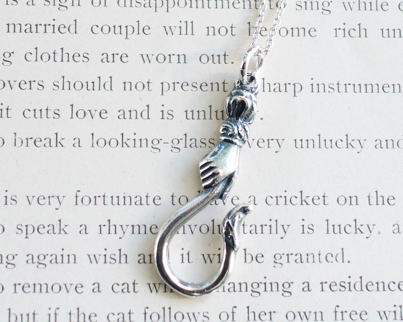 May include: A silver necklace with a charm that is a hand holding a hook. The charm is detailed with a crown and other decorative elements.
