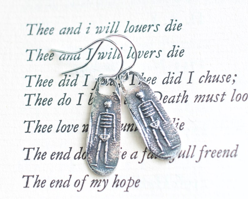 May include: Silver skeleton earrings with a textured finish. The earrings are shaped like small rectangular pendants and feature a simple skeleton design. The text "Thee did I chuse; Death must loo" is visible in the background.