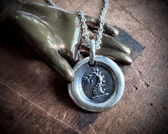 May include: A silver pendant necklace with a circular charm, featuring a detailed relief design. The charm hangs from a delicate chain. The pendant's design depicts a stylized tree or plant. The necklace is displayed on a bronze-coloured hand.